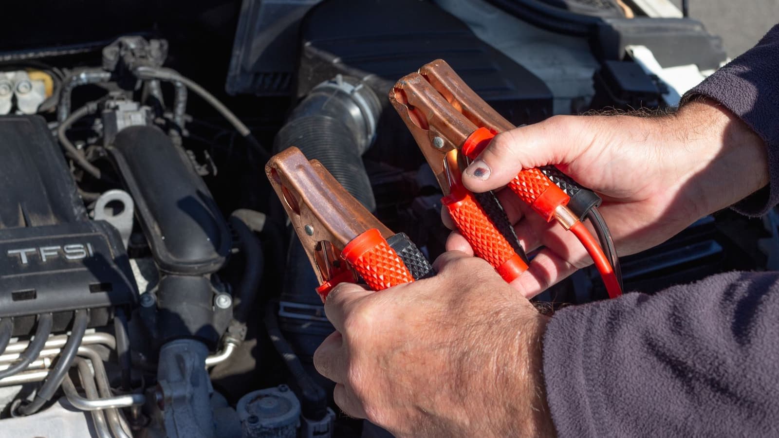 Hands holding jumper clamps in front of a car engine